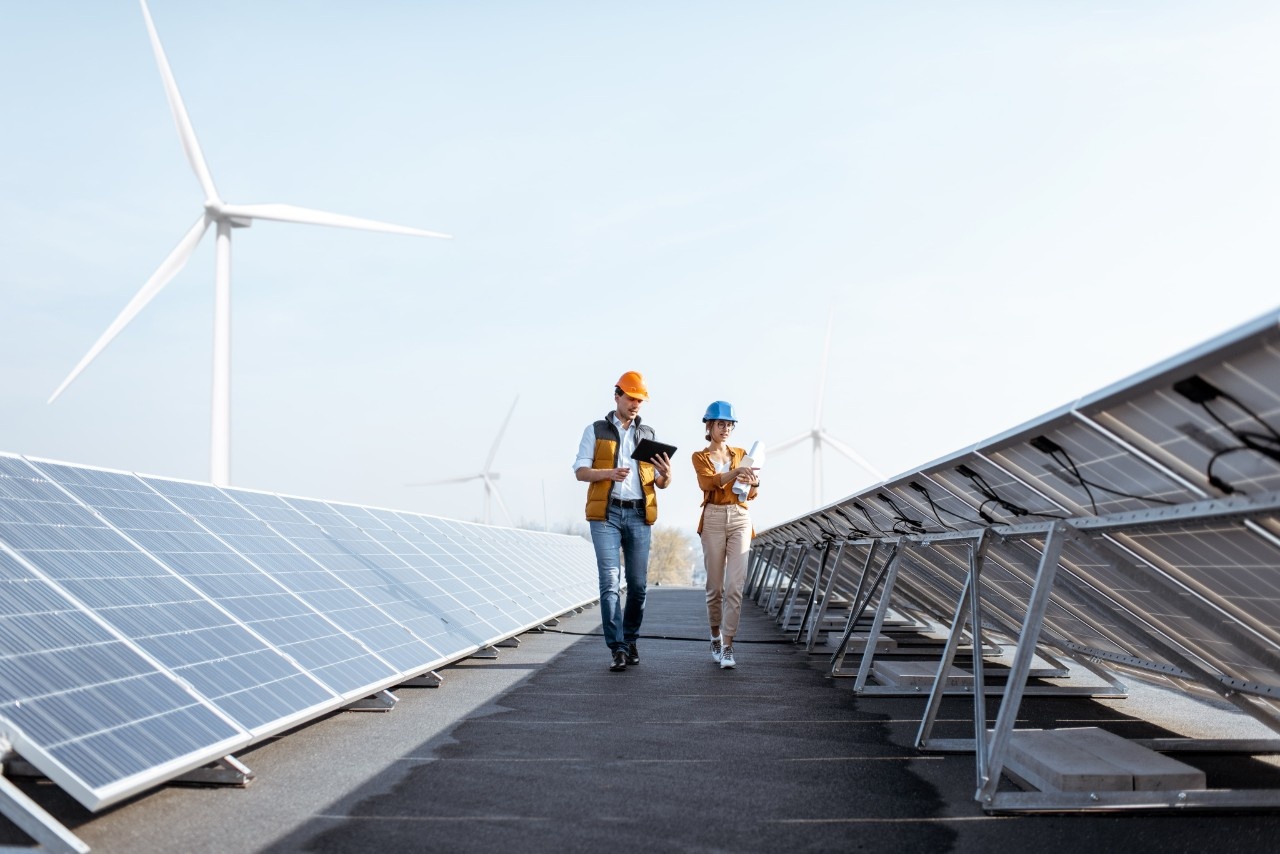 due lavoratori con caschetto e tablet in mano che camminano tra pennelli fotovoltaici, nell'atto di fare verifiche di controllo