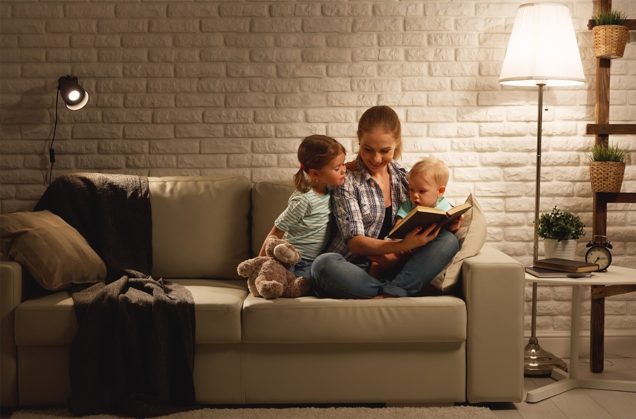 Interno di una casa, una giovane madre con due figli, un bambino biondo e una bambina poco più grande castana, su un divano bianco, che legge un libro con una luce soffusa