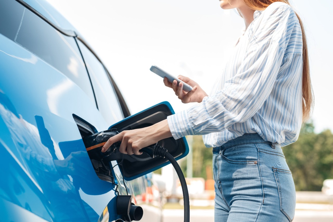 una giovane donna che fa benzina: l'inquadratura zoom sulle mani della donna che tengono la pistola della pompa di benzina e il cellulare, l'auto è azzurra e riflette l'immagine della donna; lei indossa jeans e una camicia bianca a righe azzurre