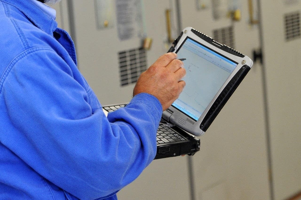 inquadratura su un computer per monitoraggio, tenuto nella mano sinistra e con la mano destra un pennino per scrivere sullo schermo, da un uomo con una camicia blu di cui non si vede il volto