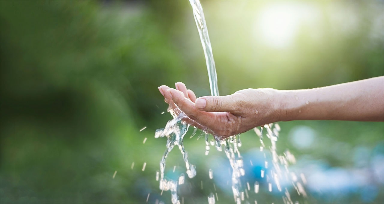 una mano tesa a raccogliere l'acqua che scende dall'alto, probabilmente da una fontanella; lo sfondo è sfocato e verde